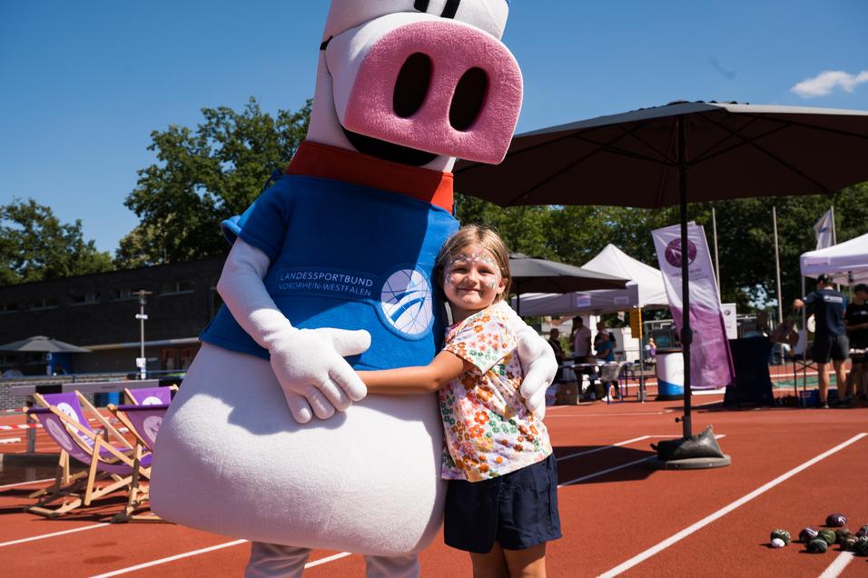 Ein Kind umarmt ein Maskottchen in Sportbekleidung auf einem Sportplatz mit Ständen und Sportequipment im Hintergrund.