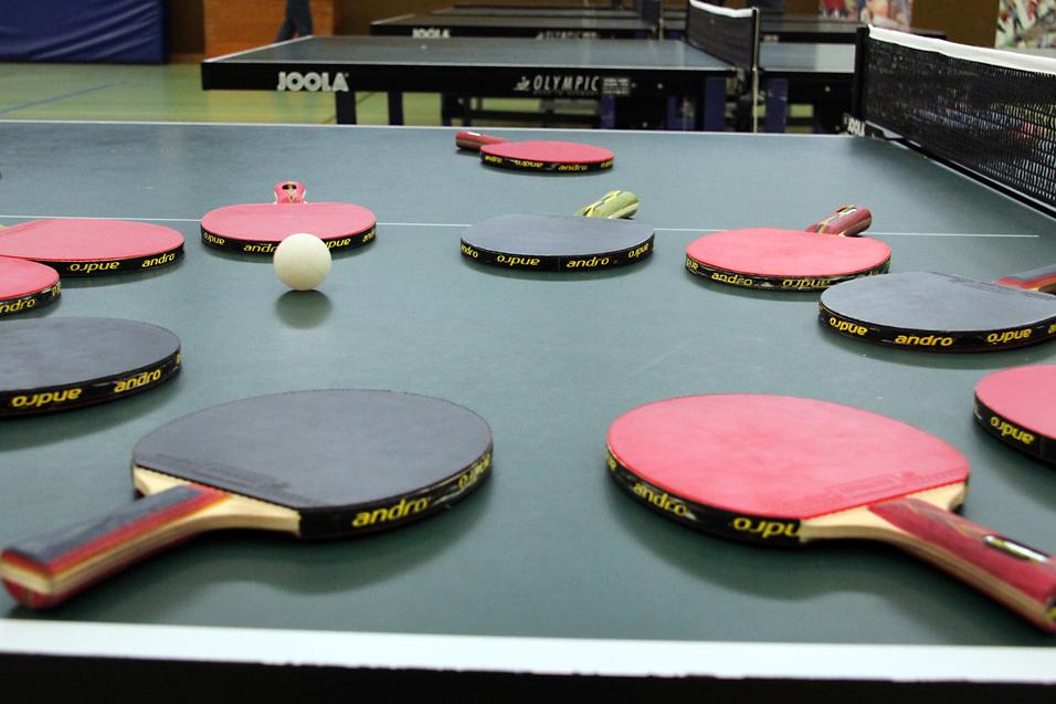 Mehrere Tischtennisschläger in verschiedenen Farben und ein Tischtennisball liegen auf einer grünen Tischtennisplatte.